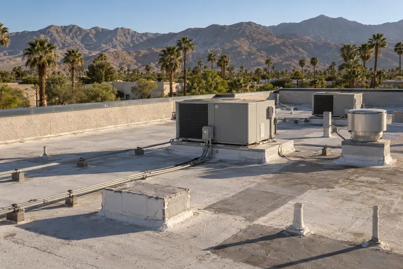 Illustrative flat commercial roof carrying packaged air-conditioning units, an exhaust fan and patches of newer coating, palms and mountains beyond