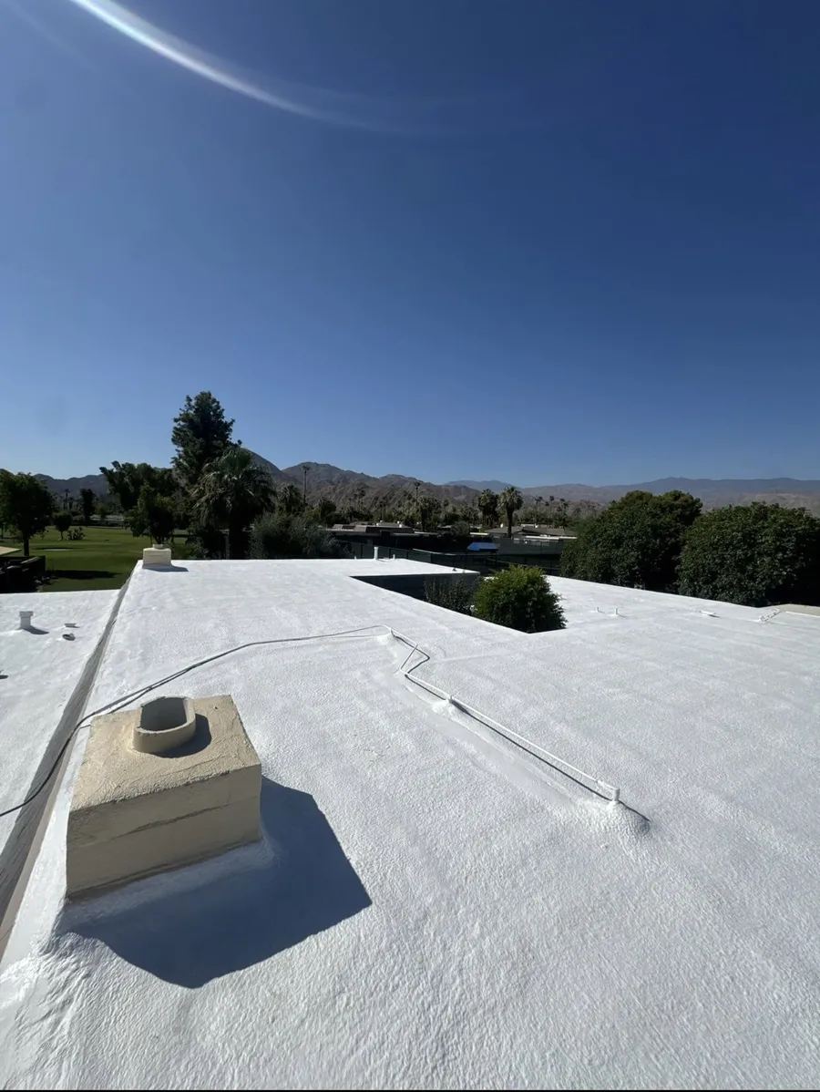 A white spray foam roof deck with a coated chimney curb and a hose run across it, mountains behind