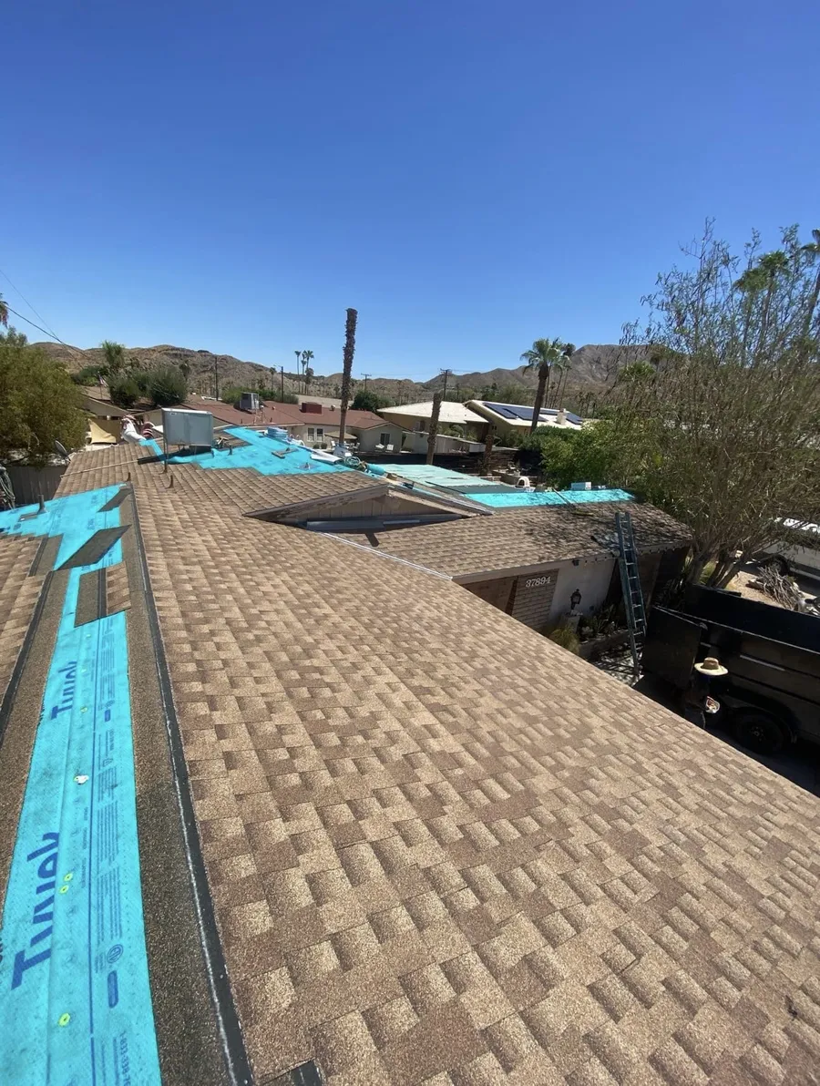 A tan shingle roof mid-installation, blue underlayment still exposed along the far edge