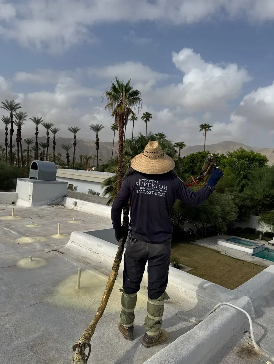 A Superior Roofing Pros crew member spraying foam across a flat roof, a pool and palms below